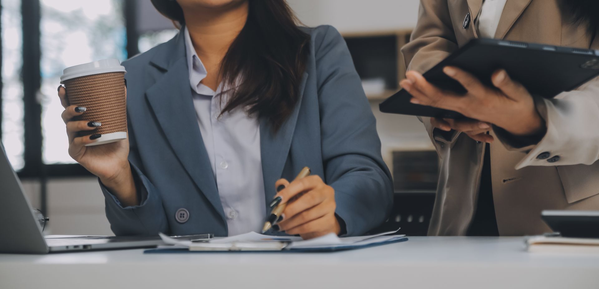 A woman is sitting at a desk with a laptop and a cup of coffee.