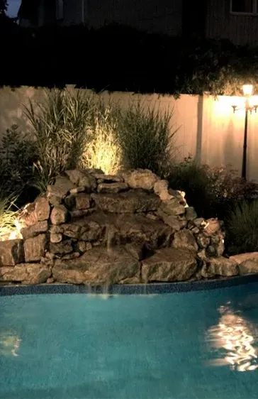 Waterfall feature with pool, lit up at night. Rocks, water, plants, and a fence are visible.