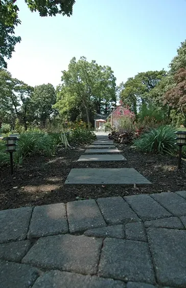 Stone pathway leading toward a gazebo surrounded by green trees and plants under a clear sky.