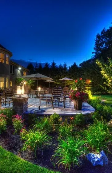 Patio at night with lit fire pit, tables, umbrellas, flowers, and trees under a dark blue sky.