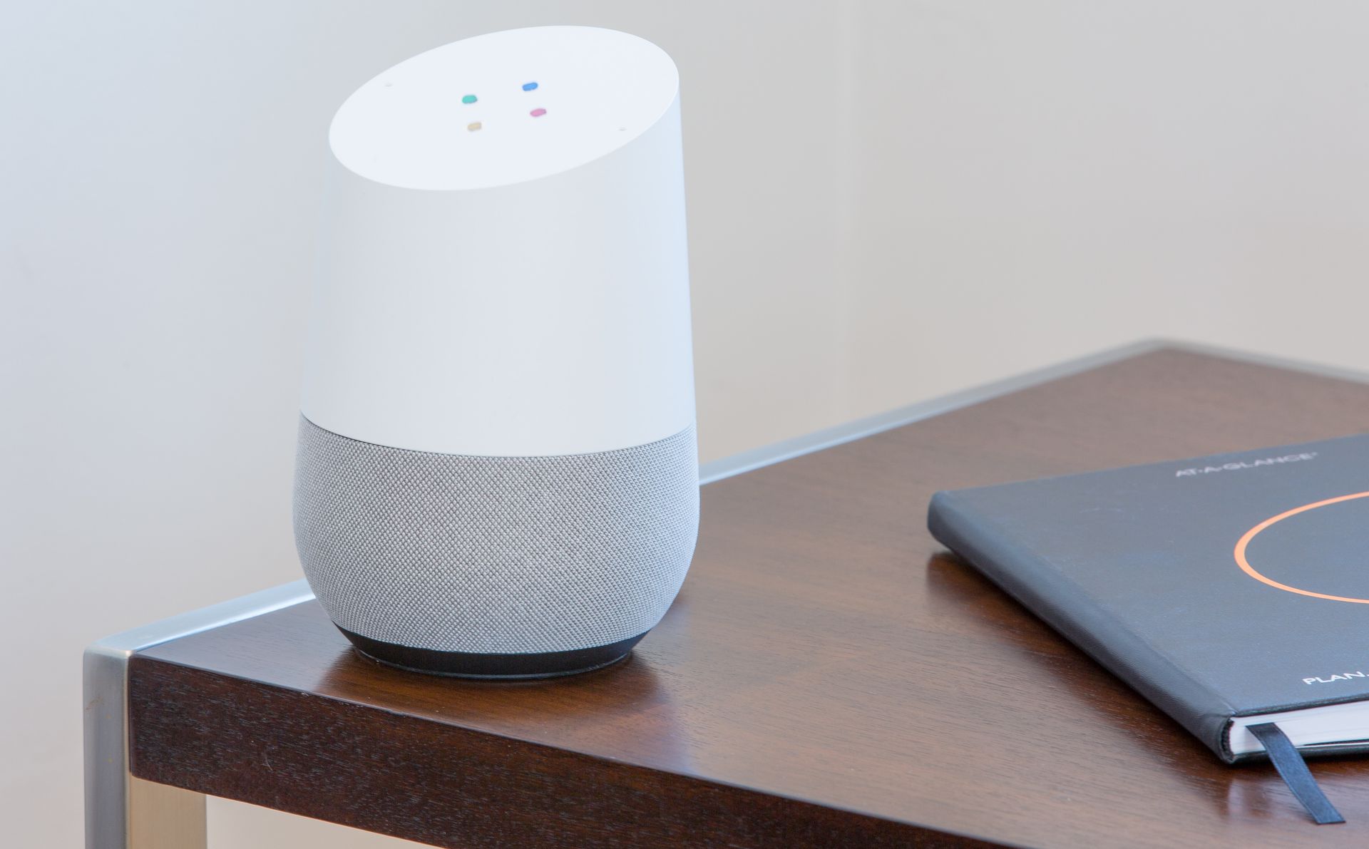 White Google Home speaker on a wooden nightstand, next to a closed notebook.