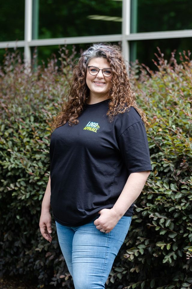 A woman wearing glasses and a black t-shirt is standing in front of a bush.