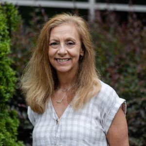 A woman in a plaid shirt is smiling for the camera while standing in front of a bush.