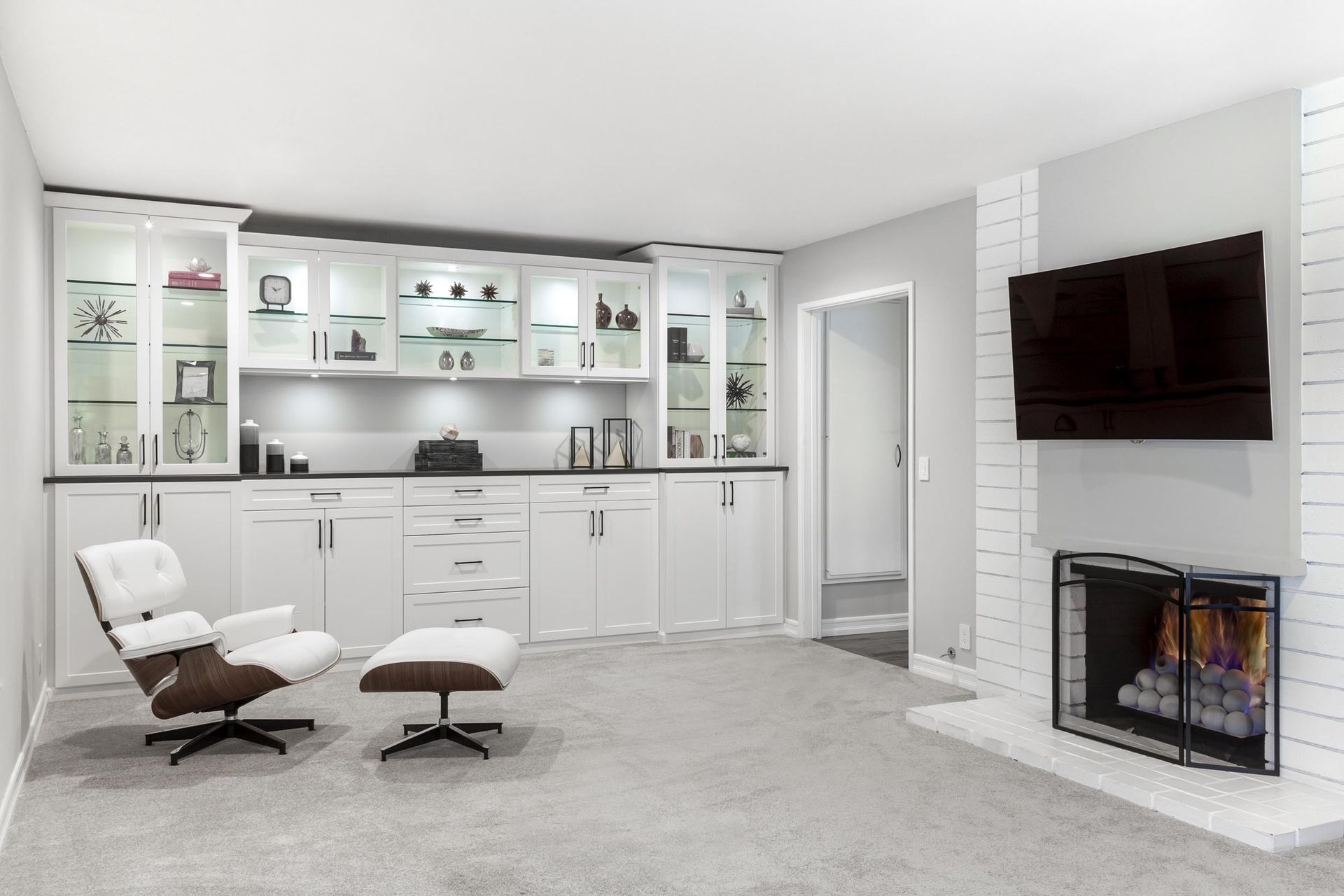 Modern living room with white built-ins, fireplace, wall-mounted TV, and a white lounge chair with ottoman.