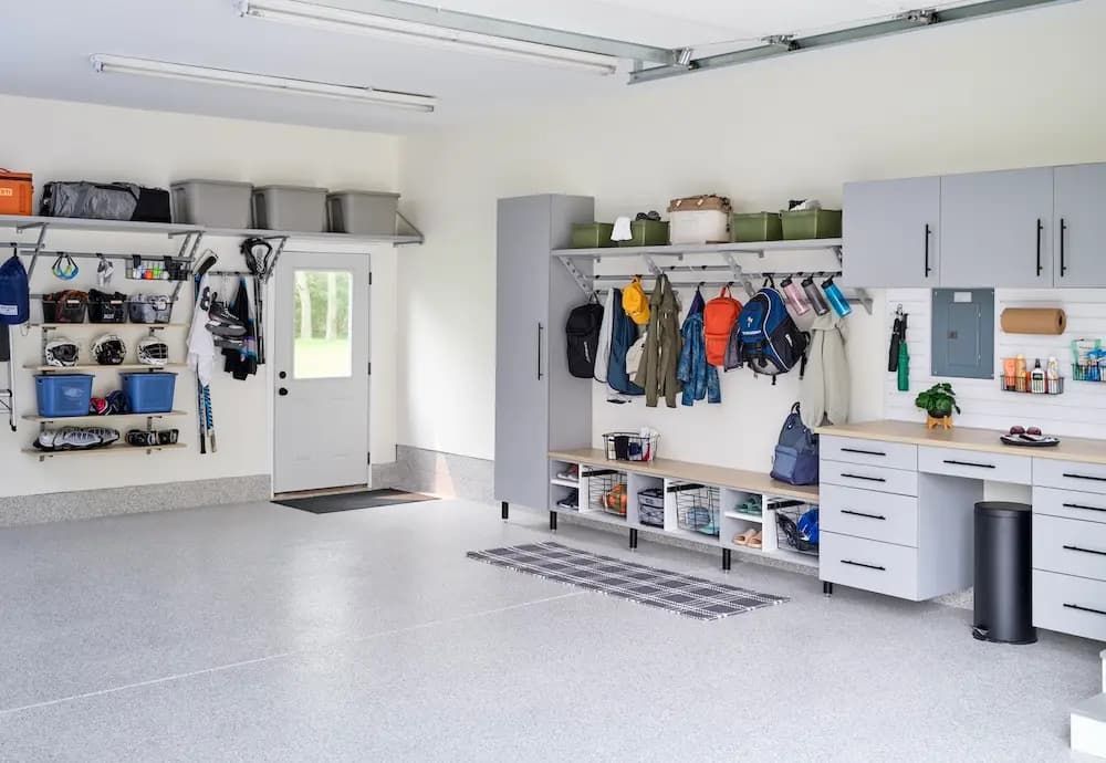 Bright garage with gray cabinets, tool storage, hanging coats, and a workbench by the door.