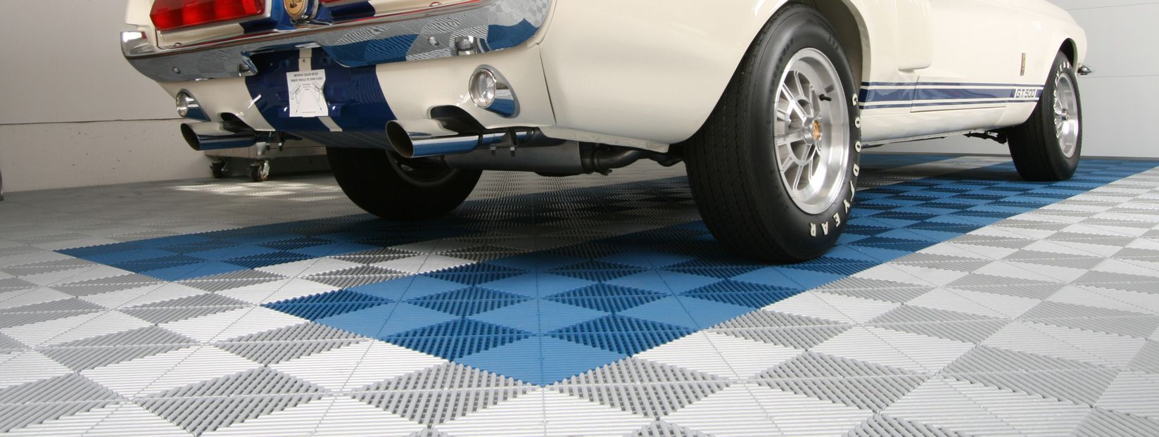 Classic white car on blue-and-white checkered floor, rear view showing chrome bumper and exhaust.