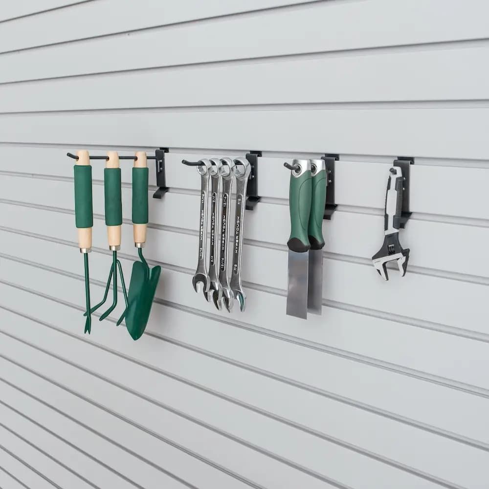 Garden tools hanging on a slat wall organizer, including green-handled trowels, wrenches, gloves, and a pruner