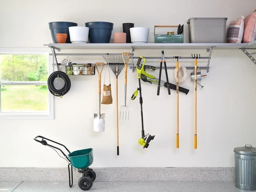 Garage wall with hanging garden tools, hoses, bins, and a wheelbarrow near a window