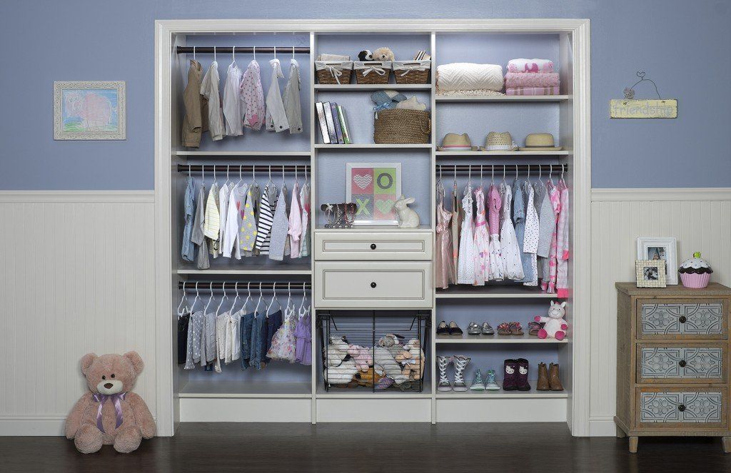 White closet of folded clothes and hanging outfits in a pastel room, with a teddy bear and small dresser.