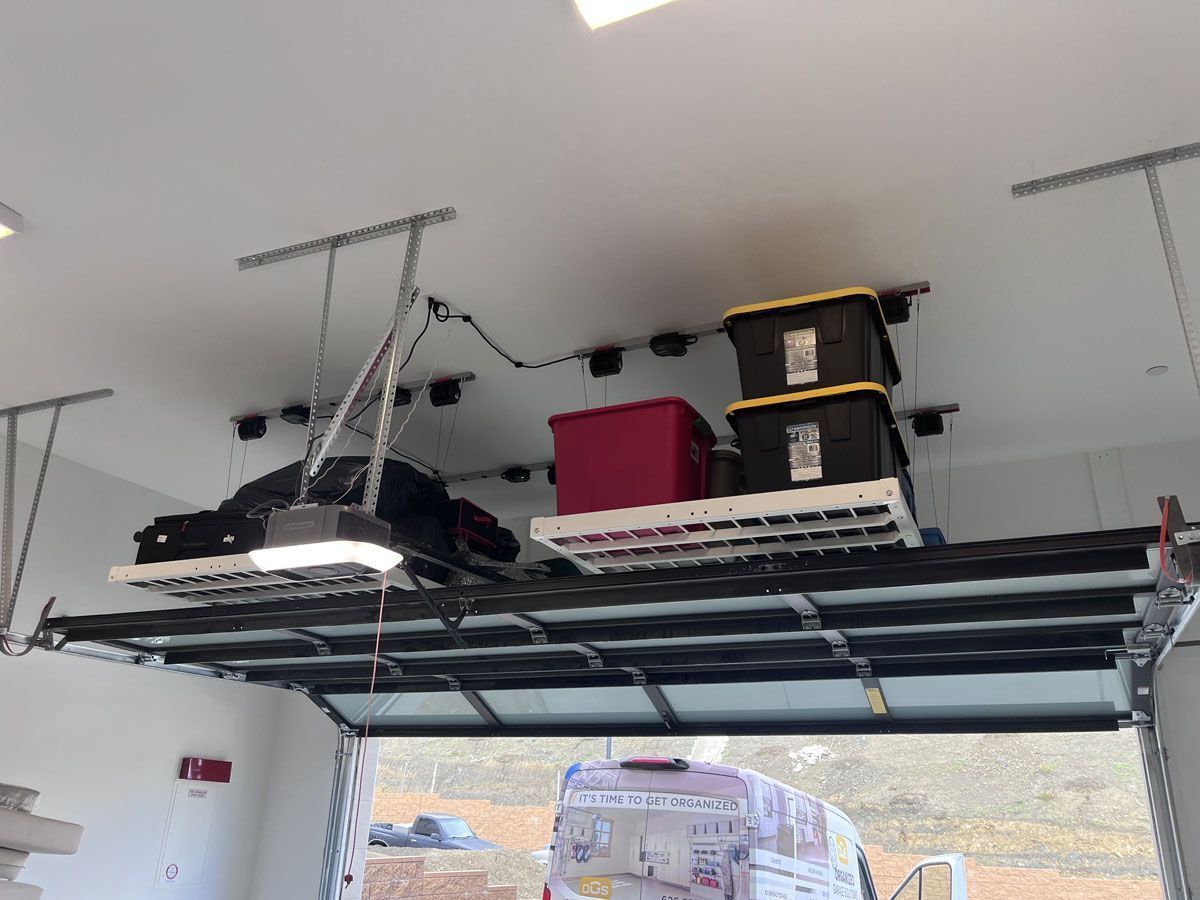 Garage ceiling storage rack holding black items, red bin, and stacked boxes above an open garage door.