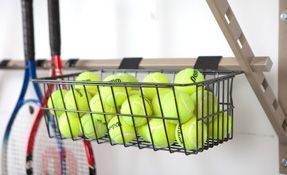 Wire basket filled with yellow tennis balls mounted on a wall next to tennis rackets and a ladder