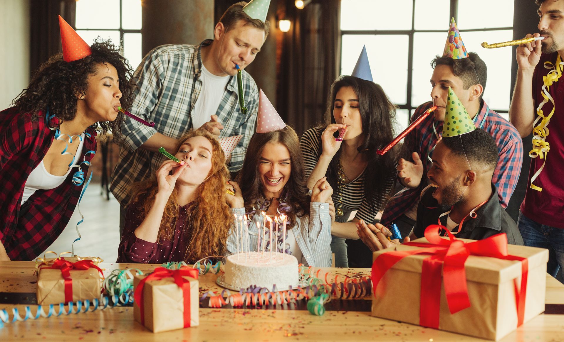 Group of people wearing party hats gathered around a birthday cake and wrapped gifts at a party.