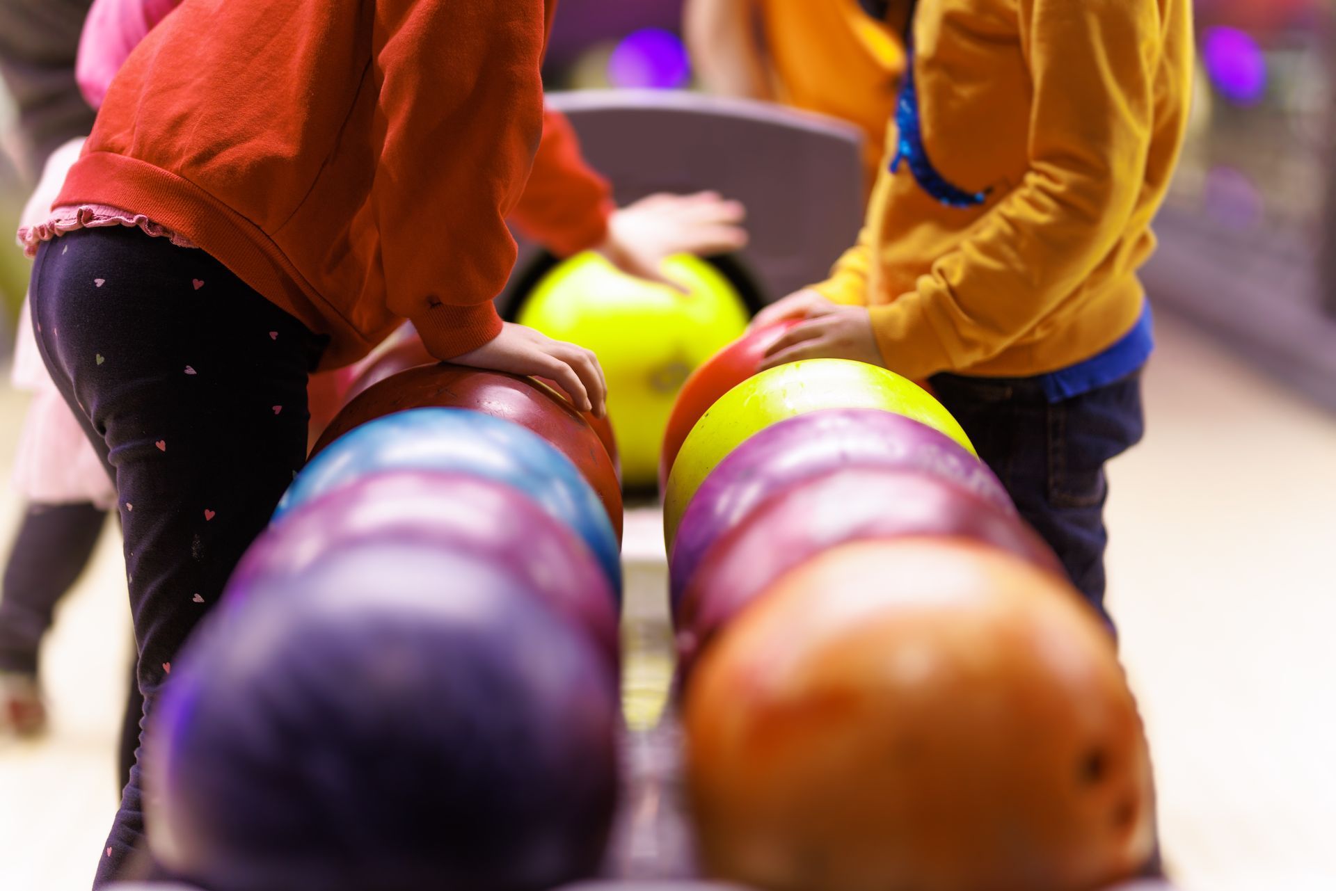 Kids reaching for colorful bowling balls on a rack at a bowling alley.