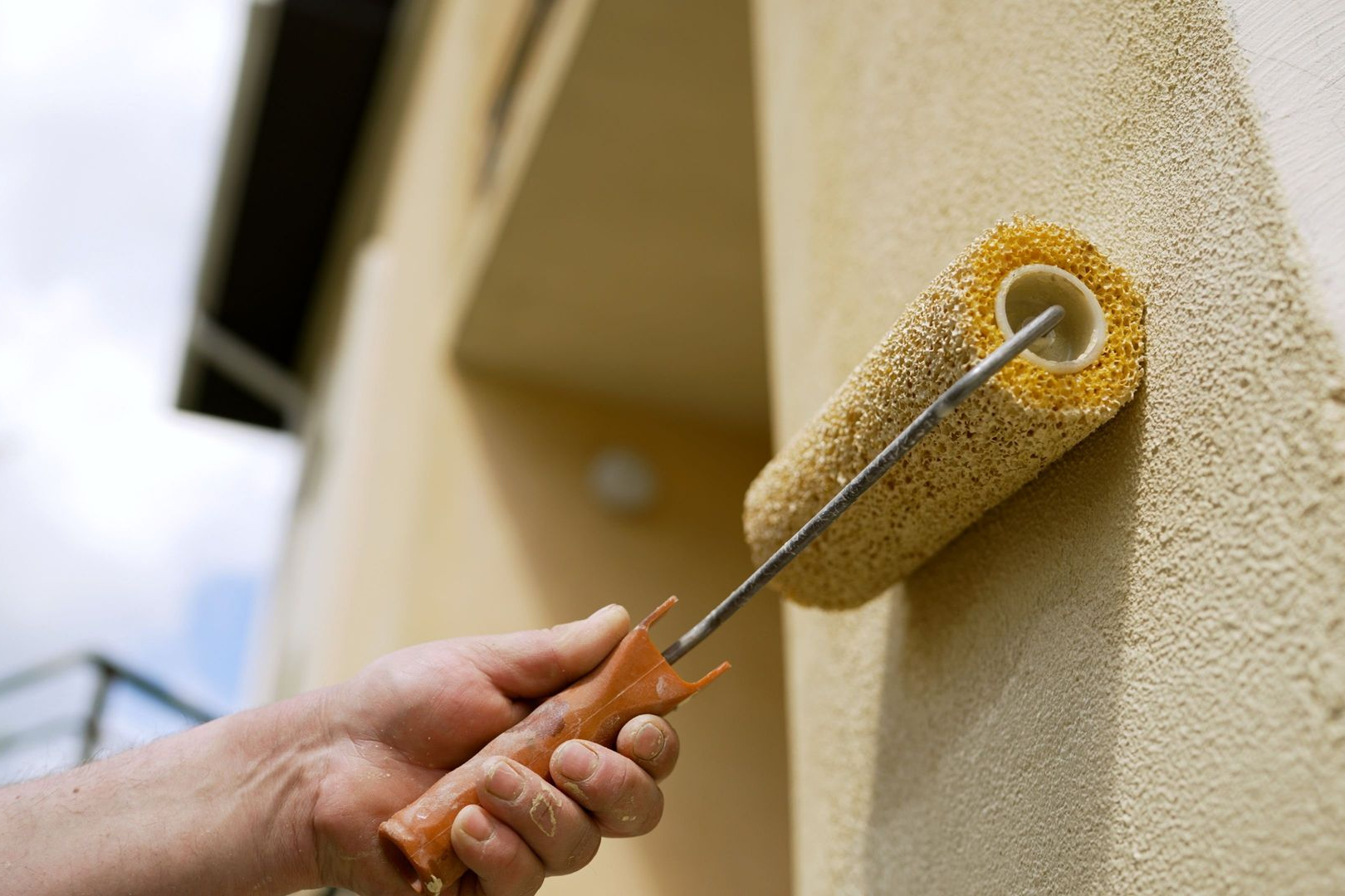 Hand using a paint roller to apply beige paint to an exterior wall