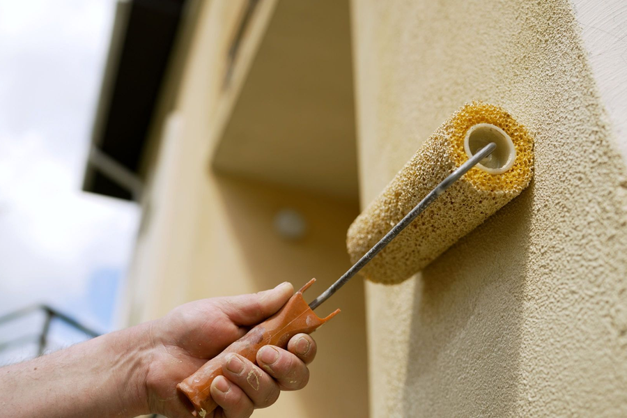Hand using a paint roller to apply beige paint to an exterior wall