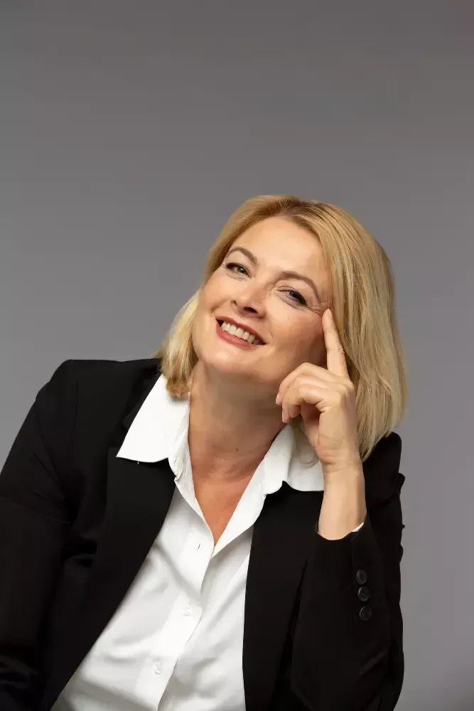 Mulher de blazer preto e camisa branca, sorrindo, tocando a testa, fundo cinza.