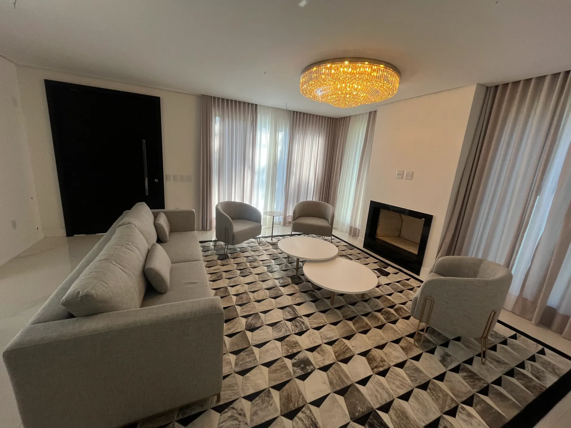 Sala de estar com sofá cinza, poltronas, tapete, lareira e lustre.