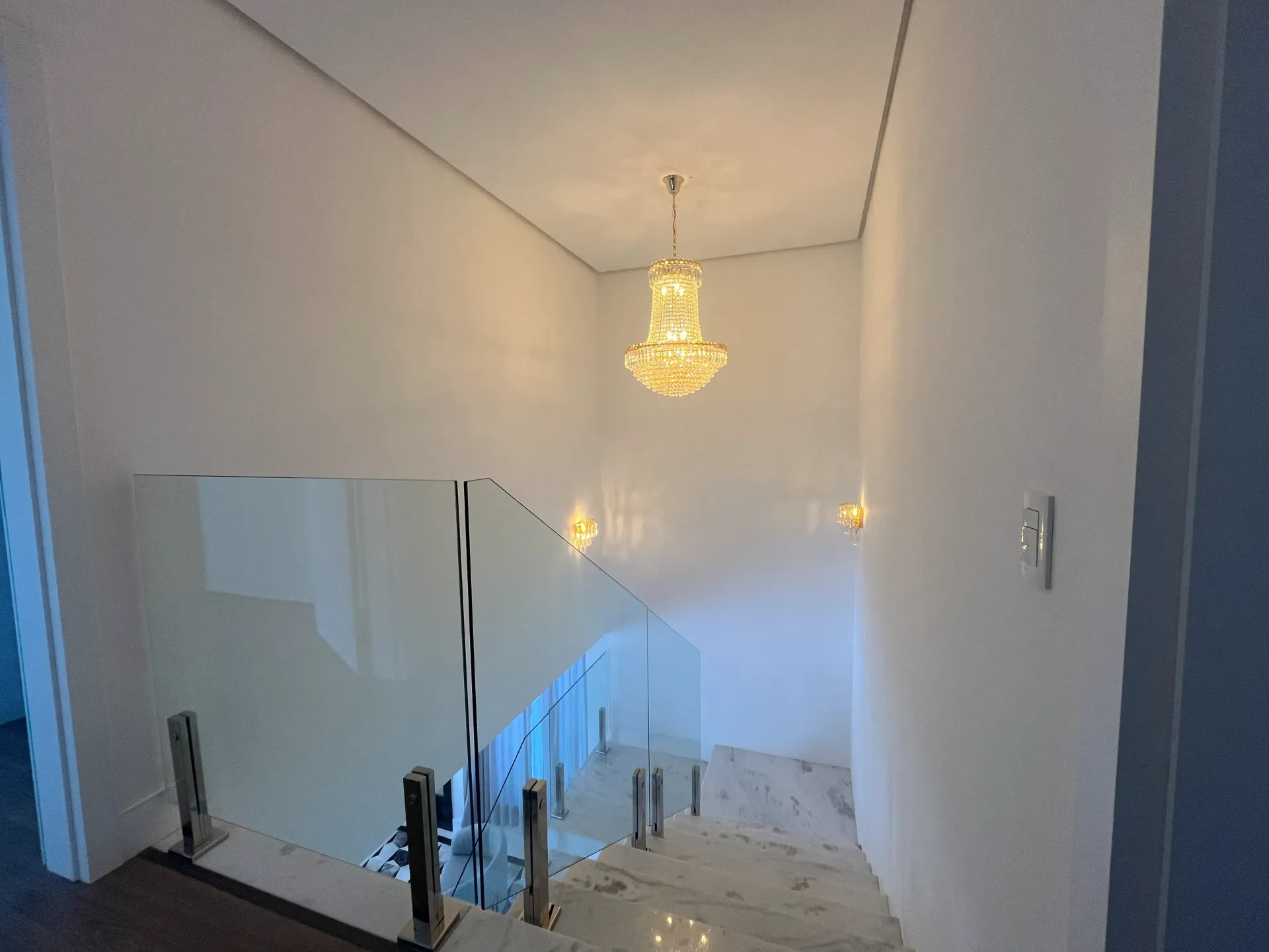 Escadaria com corrimão de vidro e lustre. Paredes brancas, piso de mármore e iluminação embutida.
