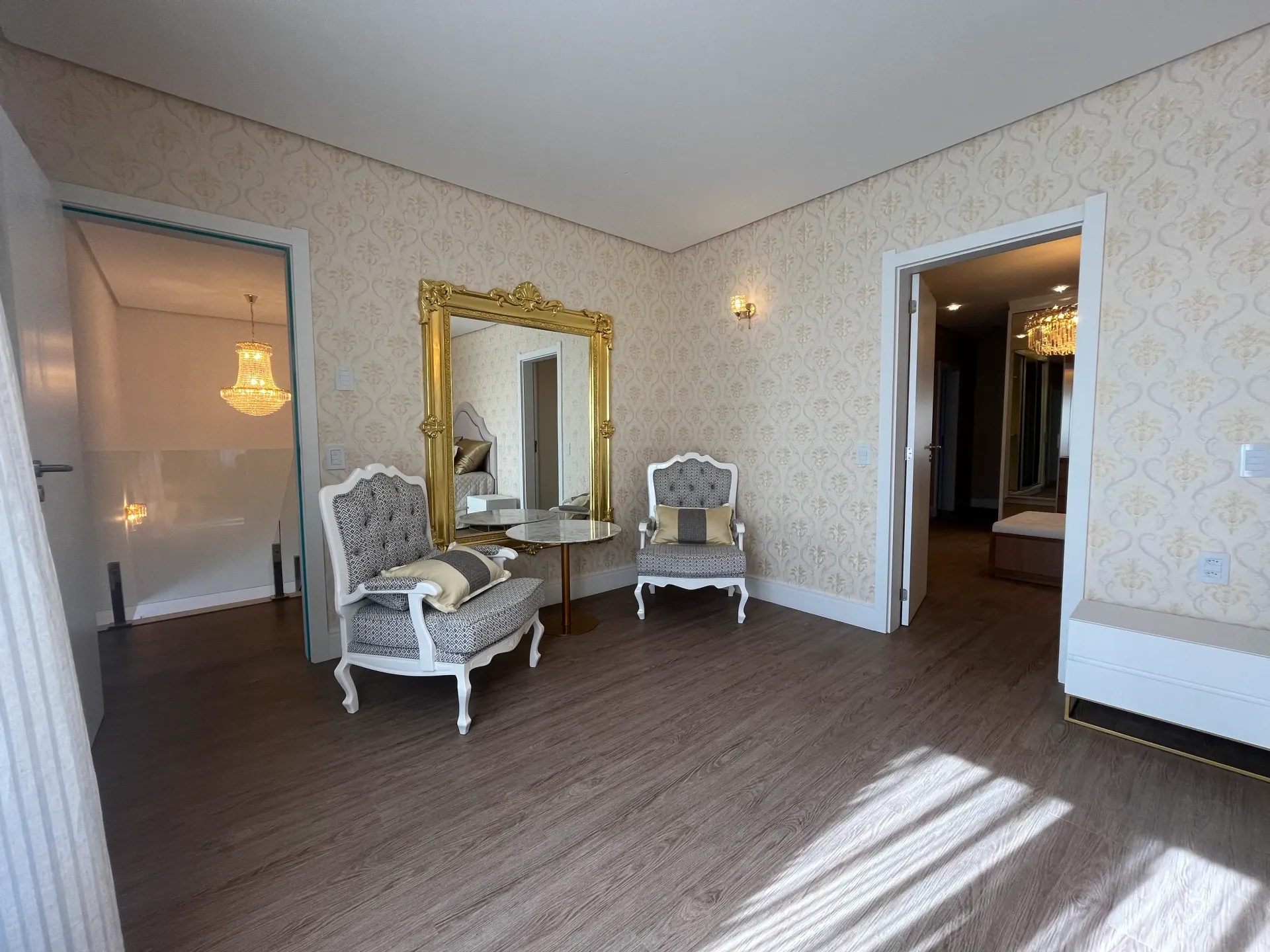 Sala de estar elegante com espelho ornamentado, poltronas e porta de acesso a um banheiro com lustre.