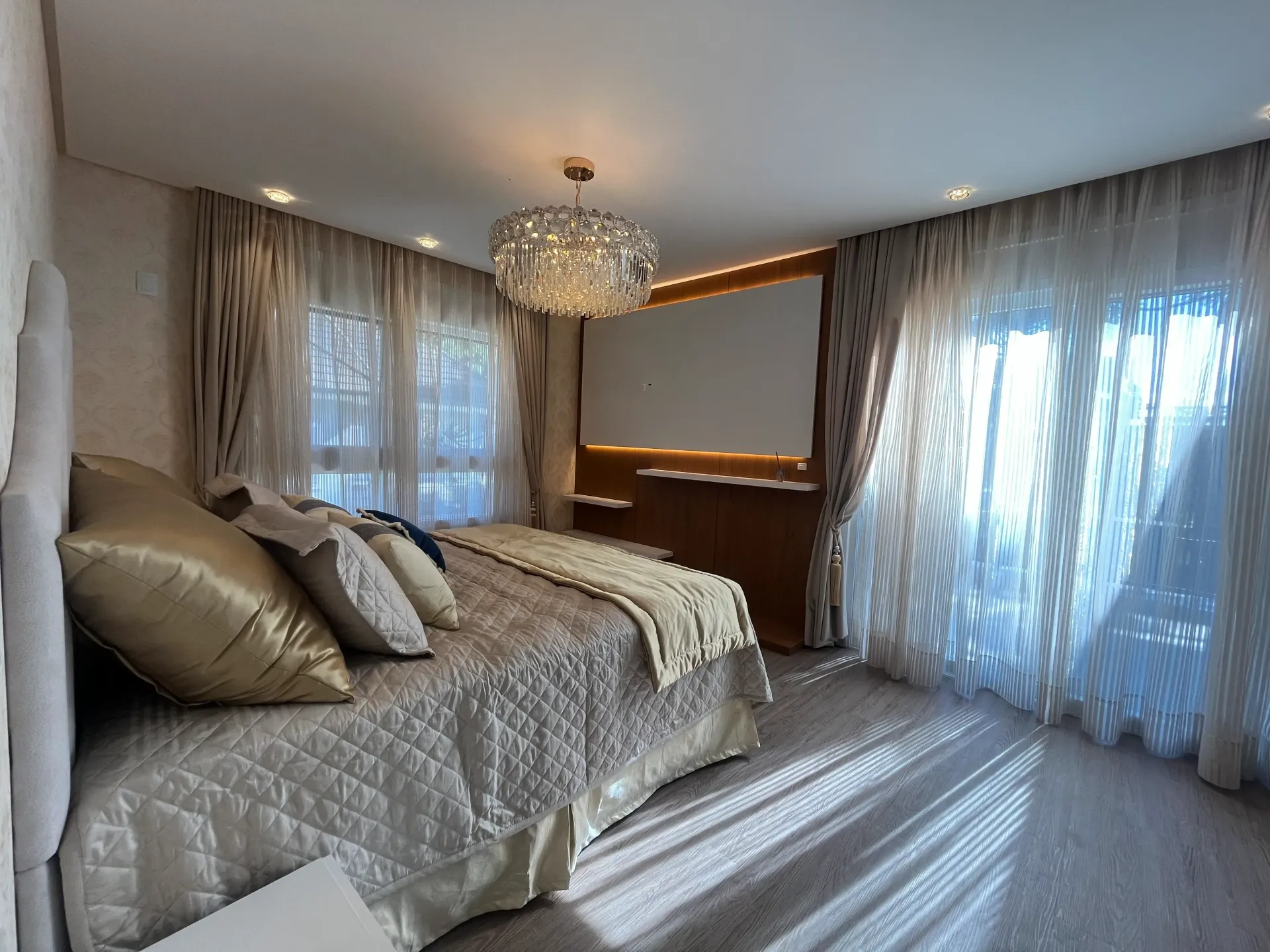 Quarto com cama, janelas amplas com cortinas translúcidas, um lustre decorativo e um armário embutido na parede.