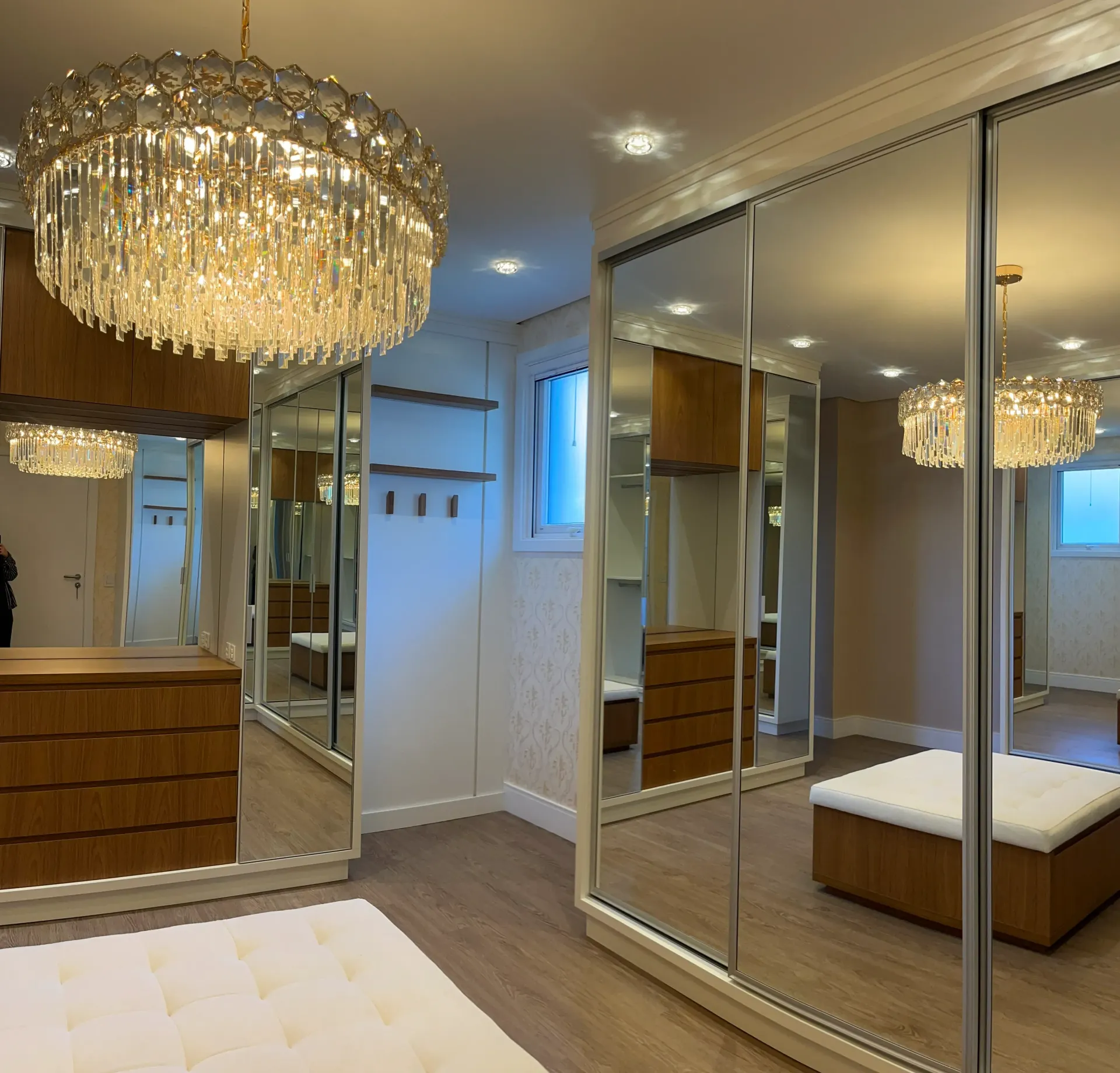 Closet com portas de correr espelhadas, lustres, cômodas e um banco. Piso e acabamentos em madeira clara.