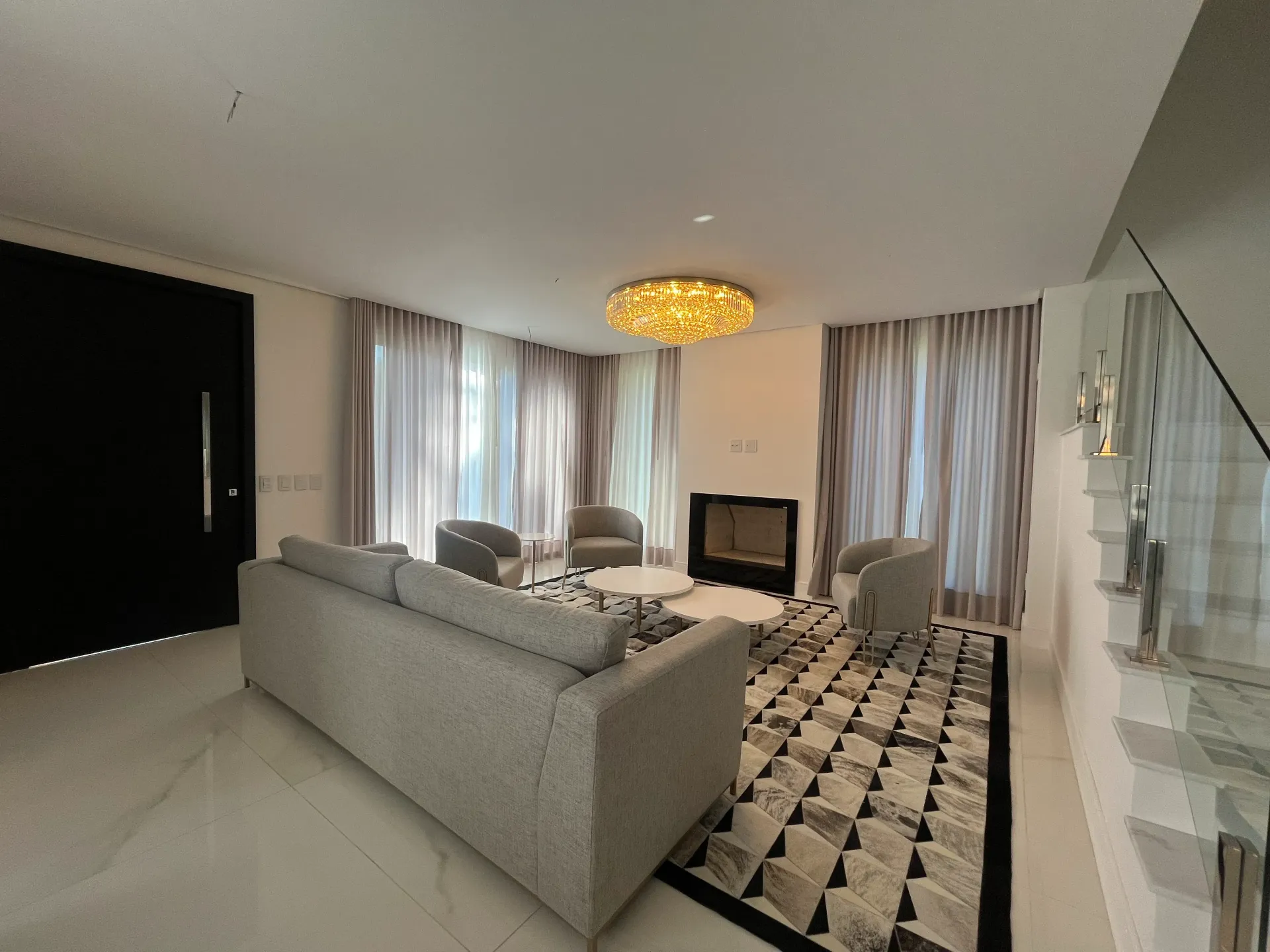 Sala de estar com sofá, cadeiras, lareira e tapete estampado; cortinas claras e luminária de teto.