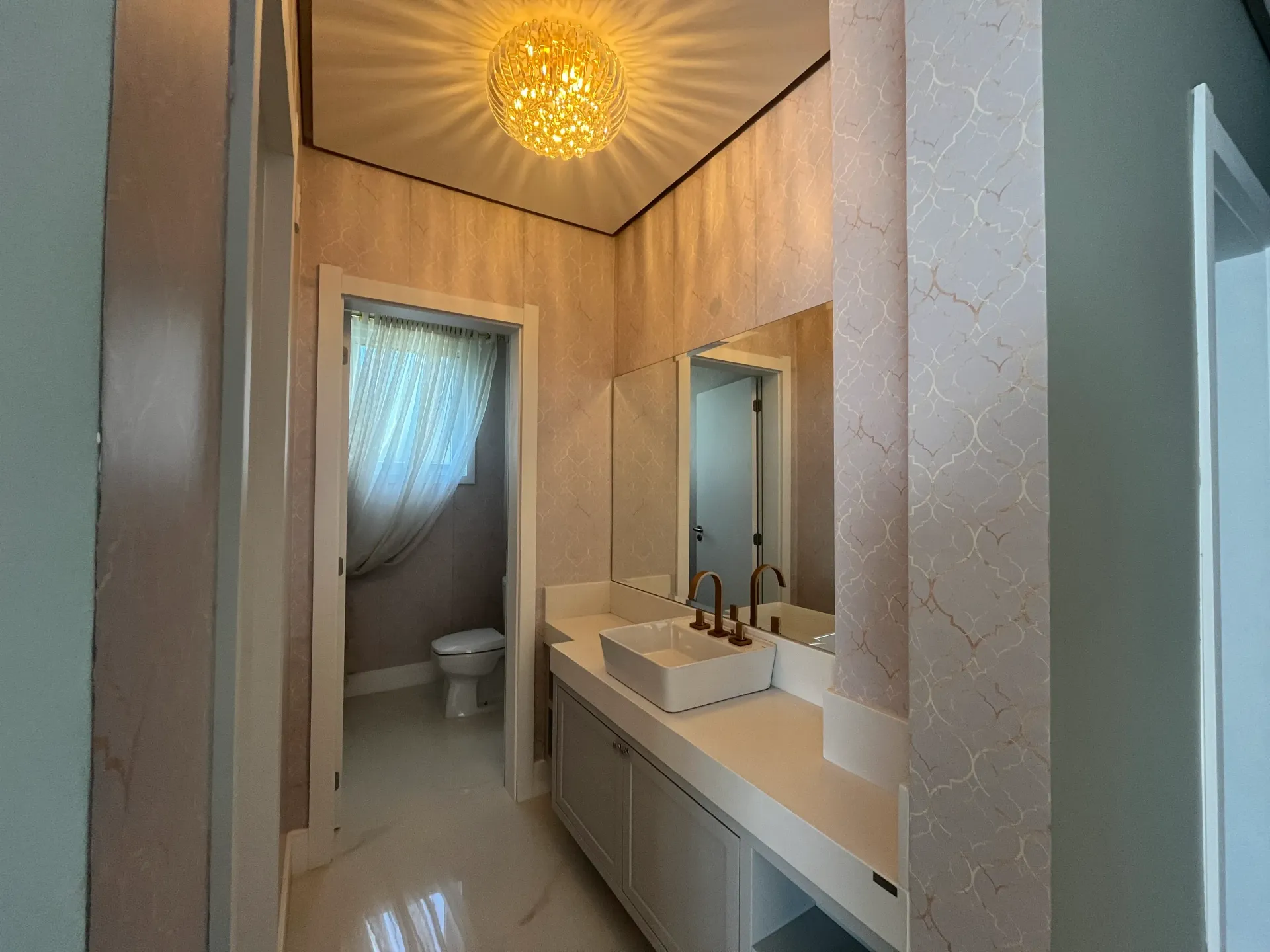 Banheiro estreito com pia, espelho e vaso sanitário brancos; papel de parede estampado, luminária pendente.