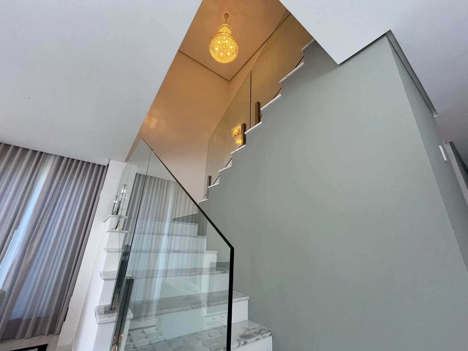 Escada com corrimão de vidro, paredes cinza e lustre.
