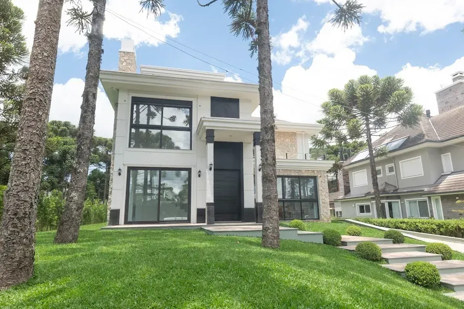 Casa branca de dois andares com grandes janelas, situada em uma colina verdejante, emoldurada por árvores.