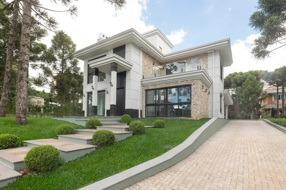 Casa moderna de dois andares com fachada de pedra e branca, gramado bem cuidado, escadaria e entrada pavimentada para carros.