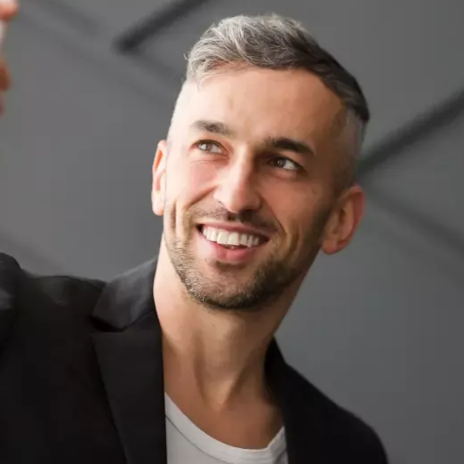Homem sorri, olha para cima, cabelo grisalho curto, jaqueta escura, camisa branca, fundo desfocado.