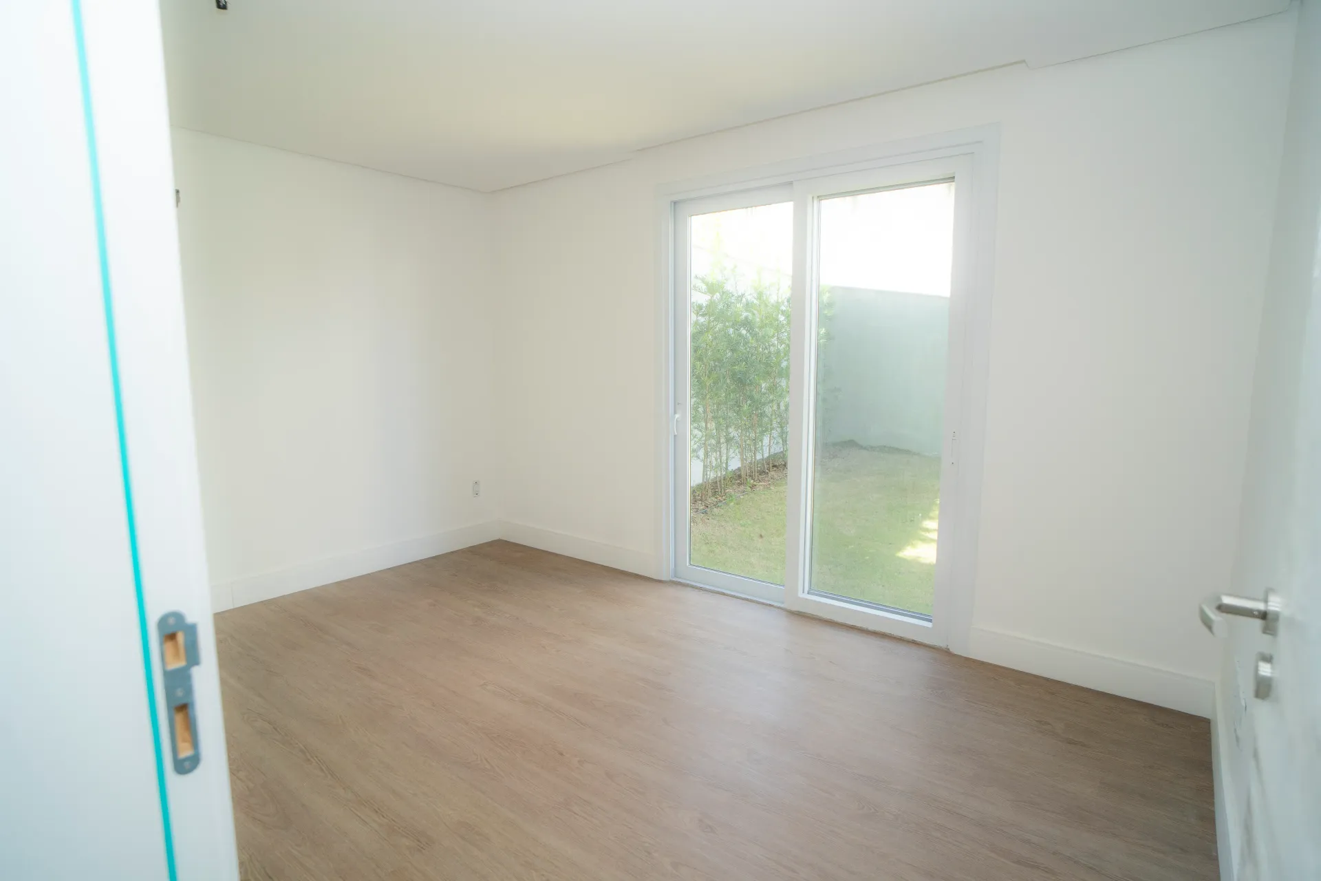 Quarto vazio com porta de vidro deslizante para um pequeno quintal. Carpete bege, paredes e portas brancas.