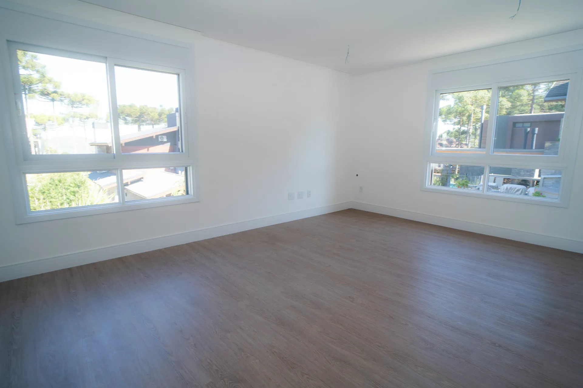 Quarto vazio com duas janelas, paredes brancas e piso que imita madeira.