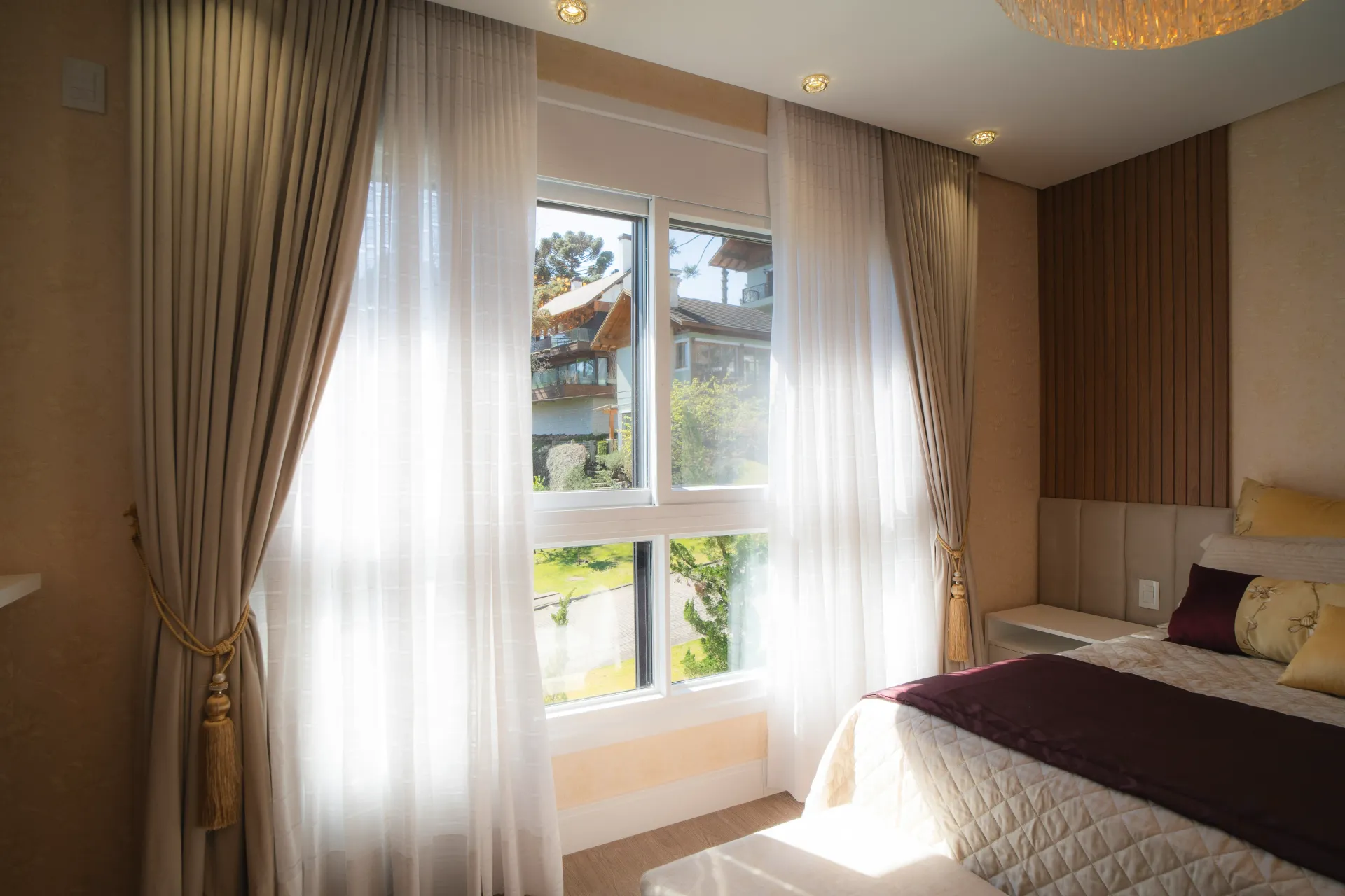Quarto com janela, cortinas brancas translúcidas, persianas bege e cama parcialmente arrumada com detalhes em tom bordô.