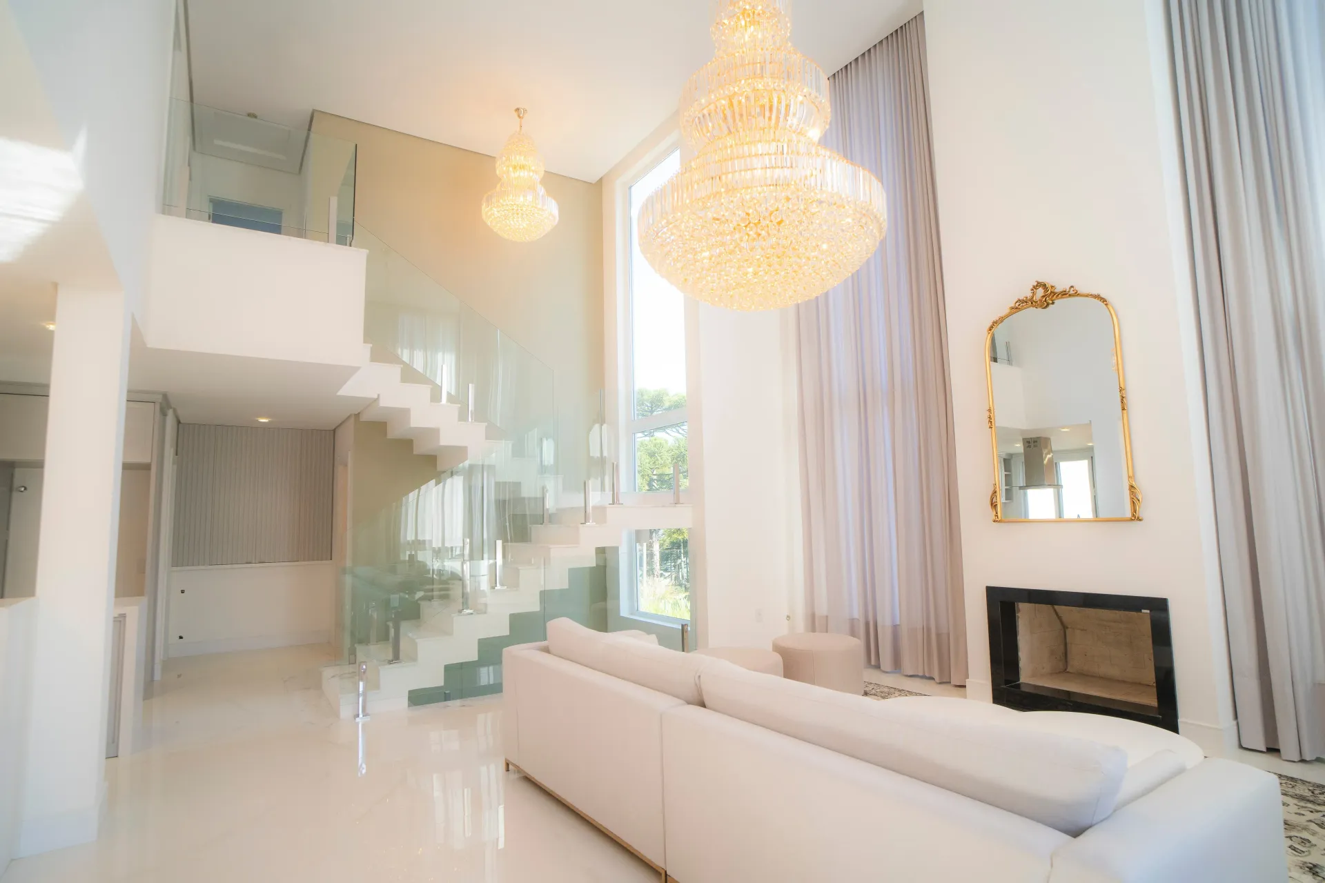 Sala de estar luxuosa em tons de branco com escada de vidro, lustre e um grande espelho com moldura dourada.