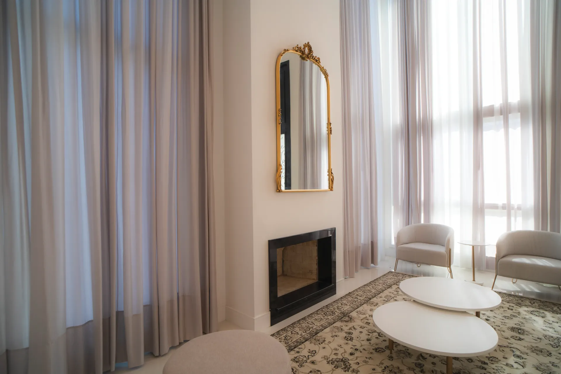 Sala de estar elegante com cortinas translúcidas, lareira, espelho dourado e mobiliário moderno.