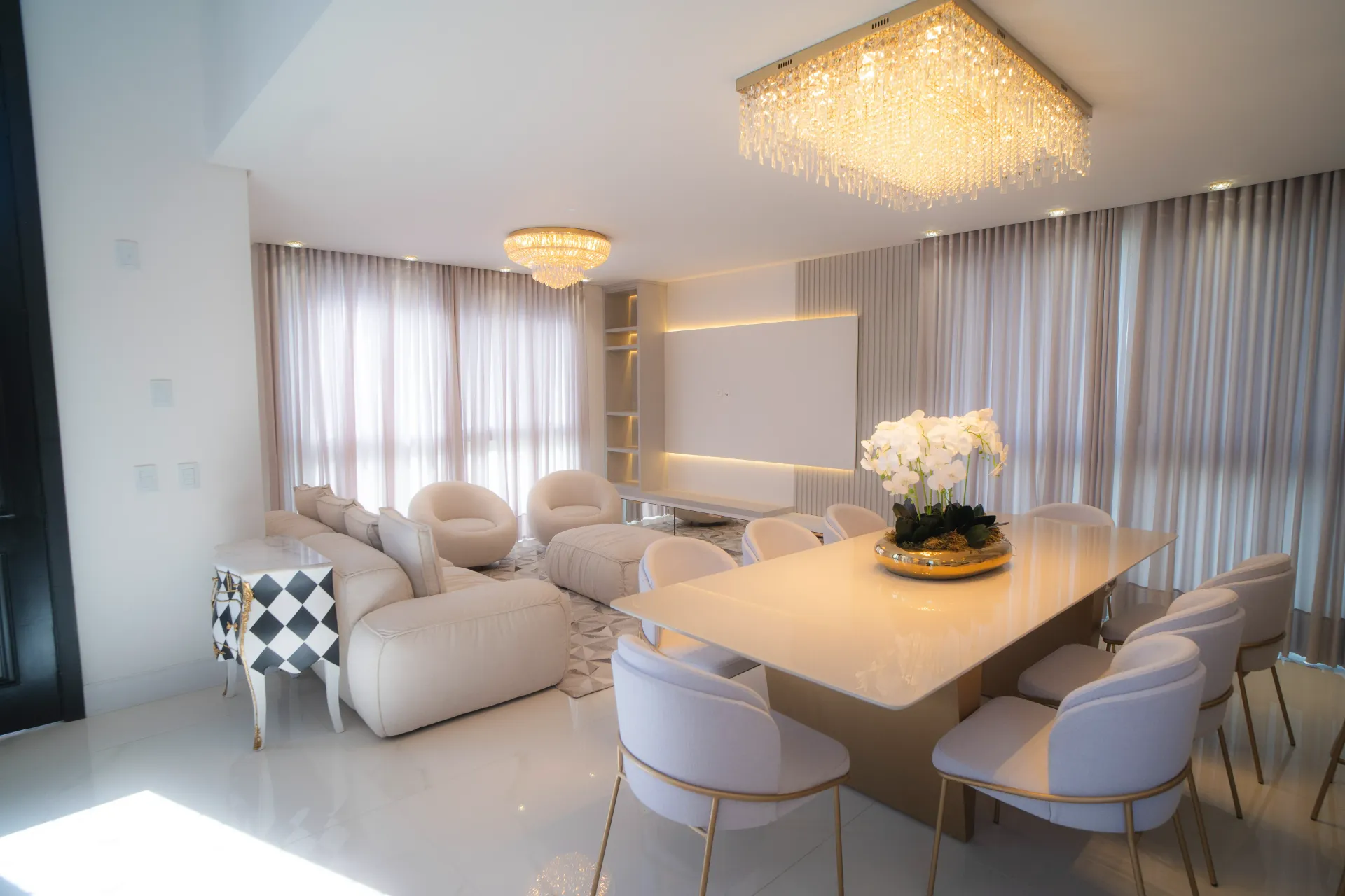 Sala de estar luxuosa com móveis em tons creme, iluminação com lustre e conjunto de mesa e jantar.