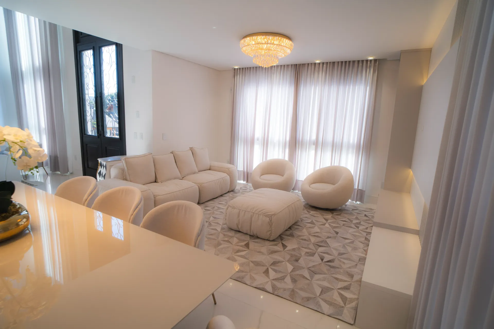 Uma sala de estar moderna com sofá, cadeiras e tapete brancos, iluminada por um lustre.