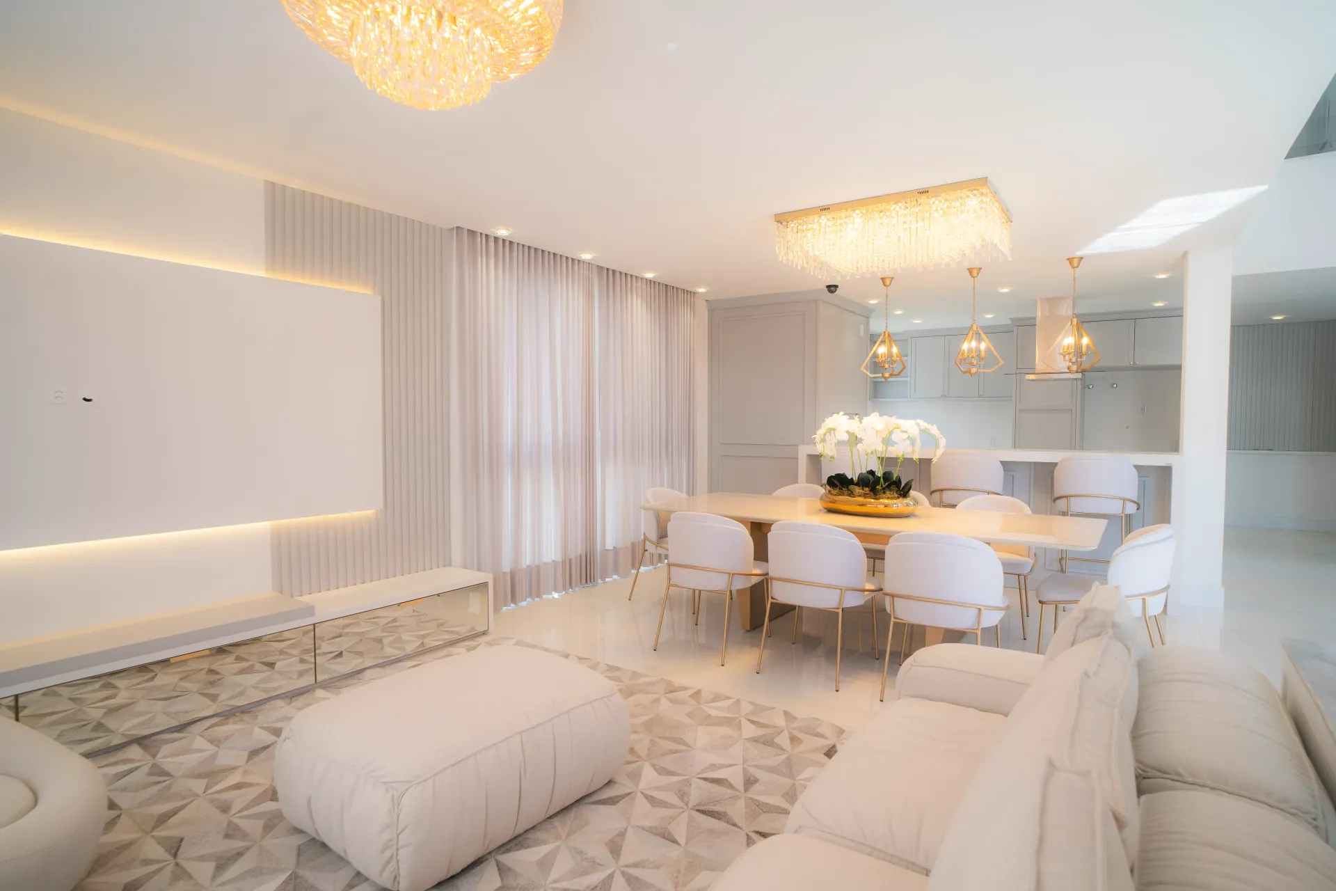 Sala de jantar e estar elegante, toda em branco, com lustres de cristal e uma longa mesa posta para receber convidados.