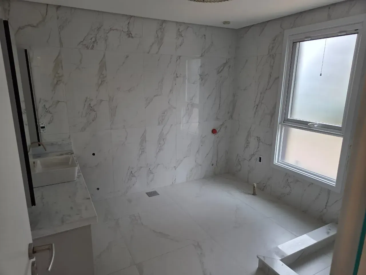 Banheiro vazio com azulejos imitando mármore, uma janela e instalações inacabadas.