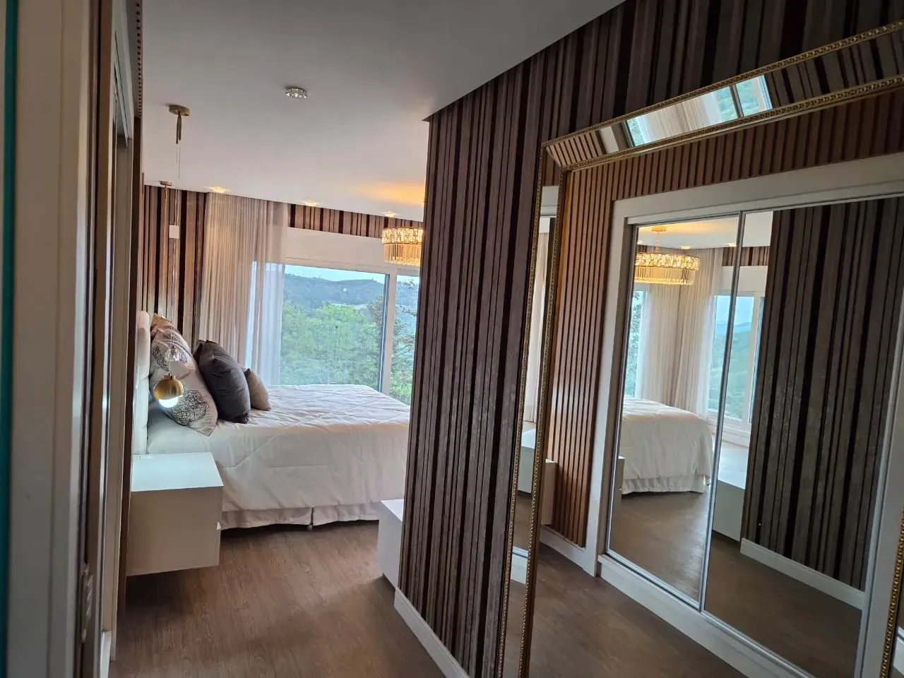 Corredor que dá para o quarto com papel de parede estampado, espelho grande e vista para a cama com lençóis brancos.