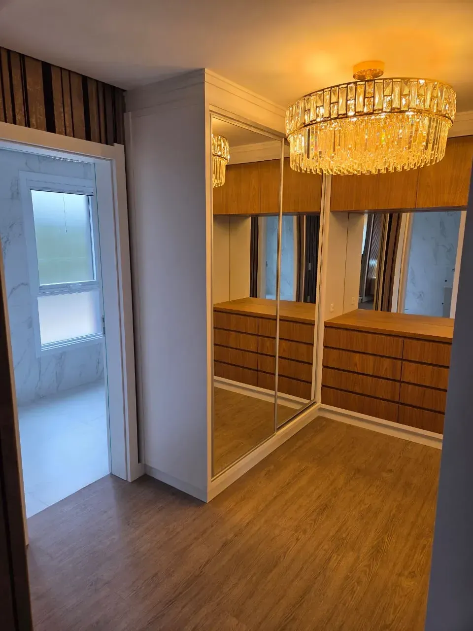Closet com portas espelhadas, gavetas em tom de madeira, lustre e vista para o corredor.