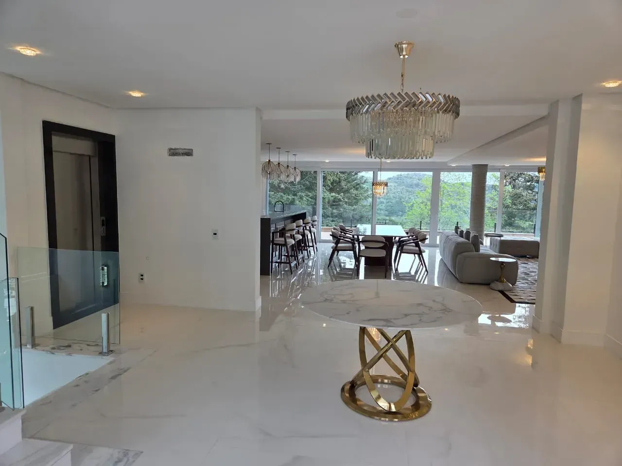 Interior luminoso e branco, de conceito aberto, com mesa de mármore e lustre, que dá acesso a uma sala de jantar.