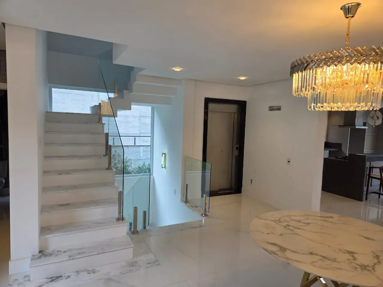 Interior luminoso e moderno com escadaria de mármore, guarda-corpo de vidro e lustre de cristal.