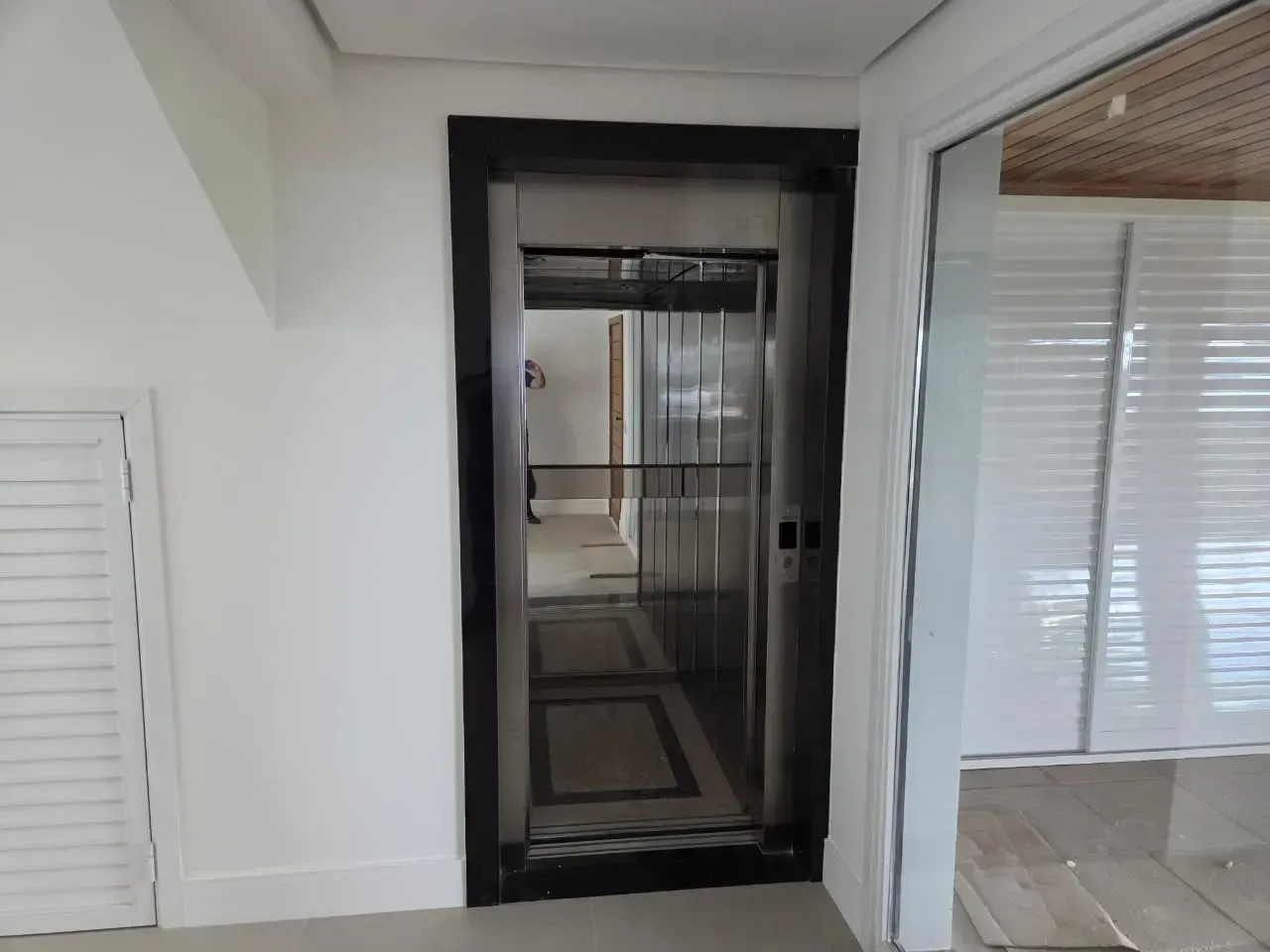 Elevador em uma sala de paredes brancas com moldura escura, interior visível.