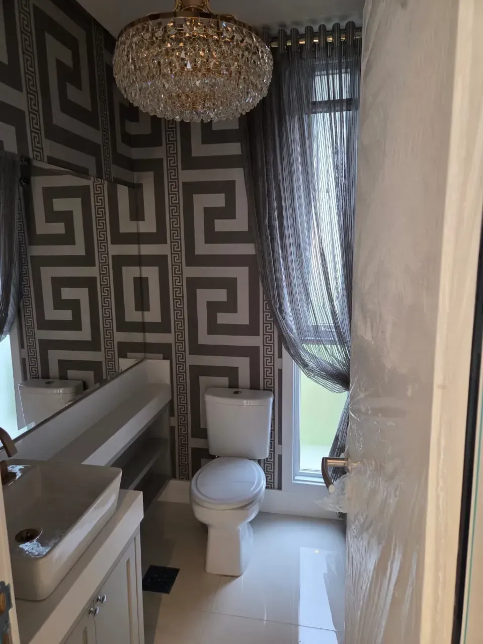 Banheiro com papel de parede geométrico cinza, lustre, vaso sanitário, pia e cortinas transparentes.