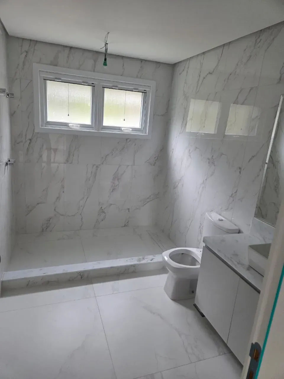 Banheiro com azulejos de mármore branco, janela, vaso sanitário e lavatório.