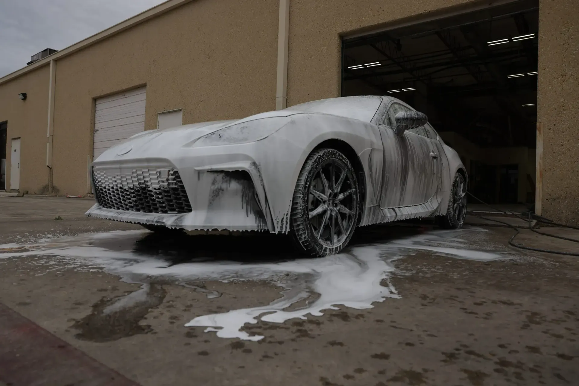 A white sports car covered in foam at a car wash, parked outside a building.