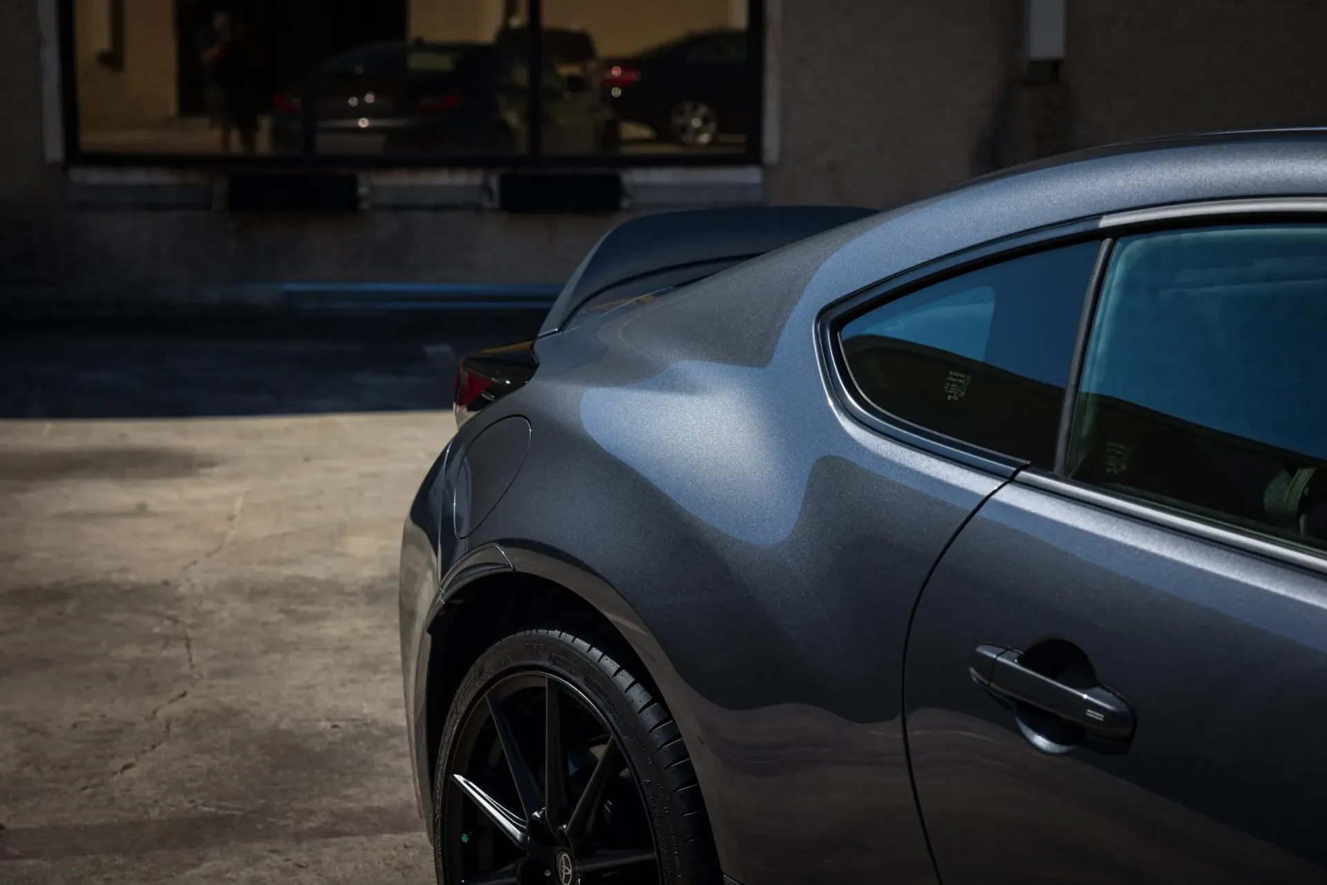 Dark gray sports car parked on concrete, with black wheels and a spoiler, reflecting the building behind it.