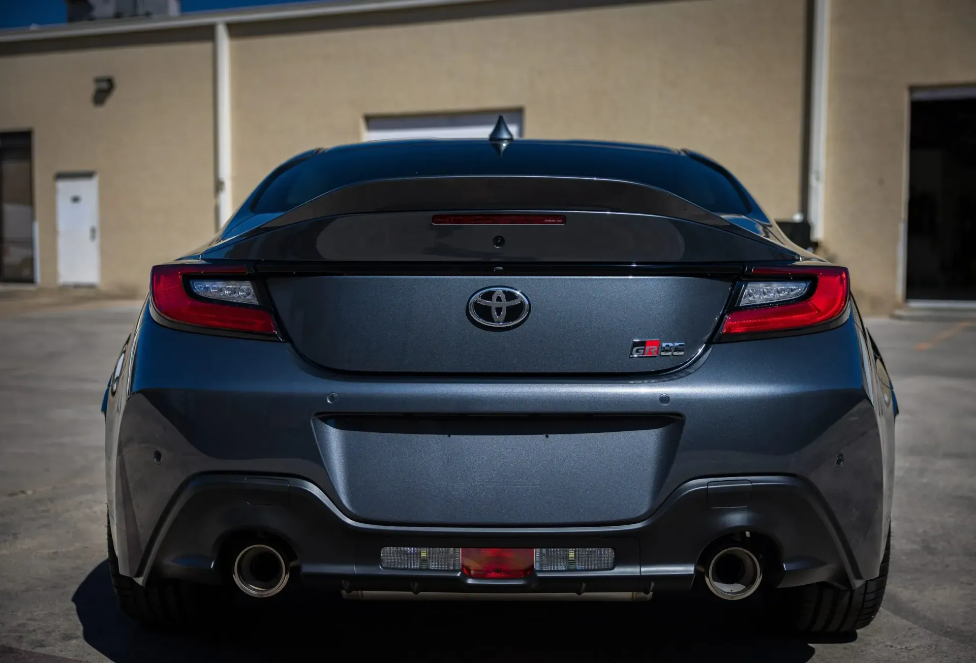 Rear view of a dark gray Toyota GR86 sports car with dual exhaust pipes, parked in front of a building.